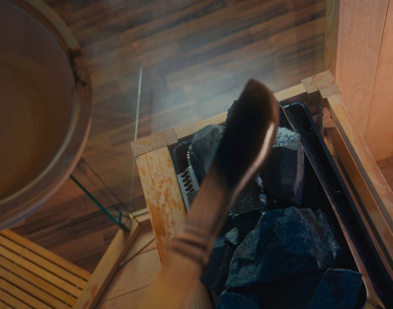 A close-up view of a sauna scene with an unidentified person using a ladle to splash water onto hot rocks, creating steam. The warm, wooden interior adds to the serene and calming atmosphere.